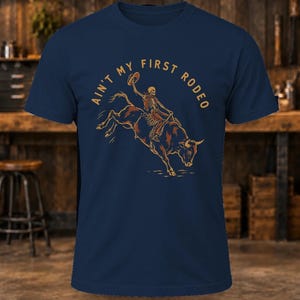 May include: A navy blue short-sleeve t-shirt featuring a graphic of a skeleton riding a bucking bull while holding a hat. The text "AIN'T MY FIRST RODEO" is printed in a tan color in an arc above the central illustration.
