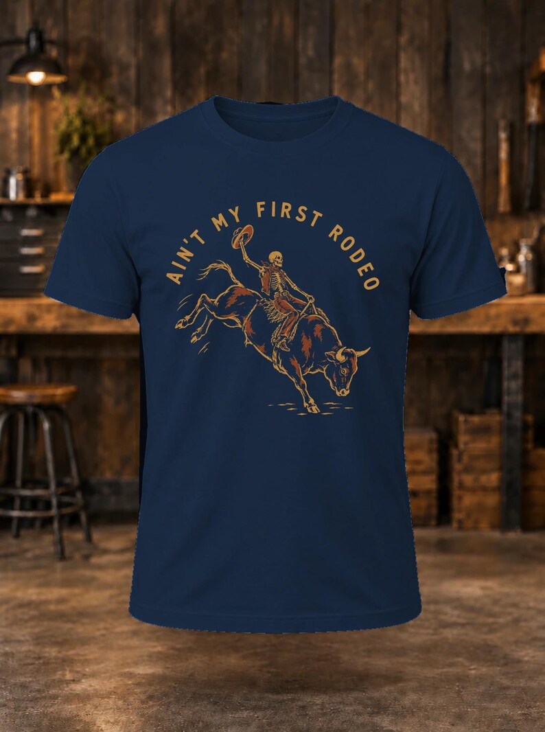 May include: A navy blue short-sleeve t-shirt featuring a graphic of a skeleton riding a bucking bull while holding a hat. The text "AIN'T MY FIRST RODEO" is printed in a tan color in an arc above the central illustration.