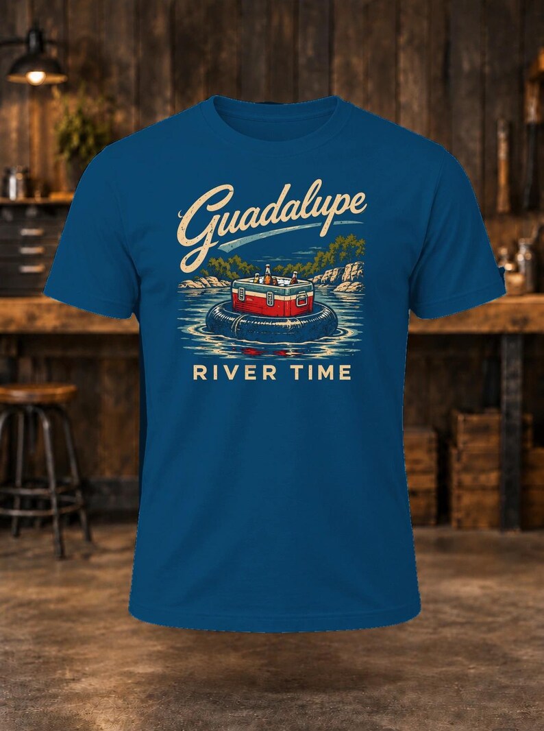 May include: A blue t-shirt featuring a graphic illustration of a red and white cooler filled with bottles, floating on a black inner tube in water. The text "Guadalupe" is printed in a script font above, and "RIVER TIME" is printed in block letters below.