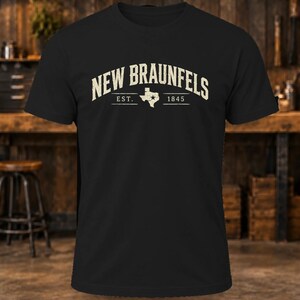 May include: A black short-sleeve t-shirt featuring the text NEW BRAUNFELS in a distressed cream font. Below the text is the abbreviation EST. and the year 1845, separated by a white silhouette of the state of Texas with a star.