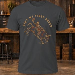 May include: A charcoal gray short-sleeve t-shirt featuring a graphic of a skeleton riding a bucking bull. The text "AIN'T MY FIRST RODEO" is printed in an arc above the skeleton in a tan color. The design is centered on the chest.