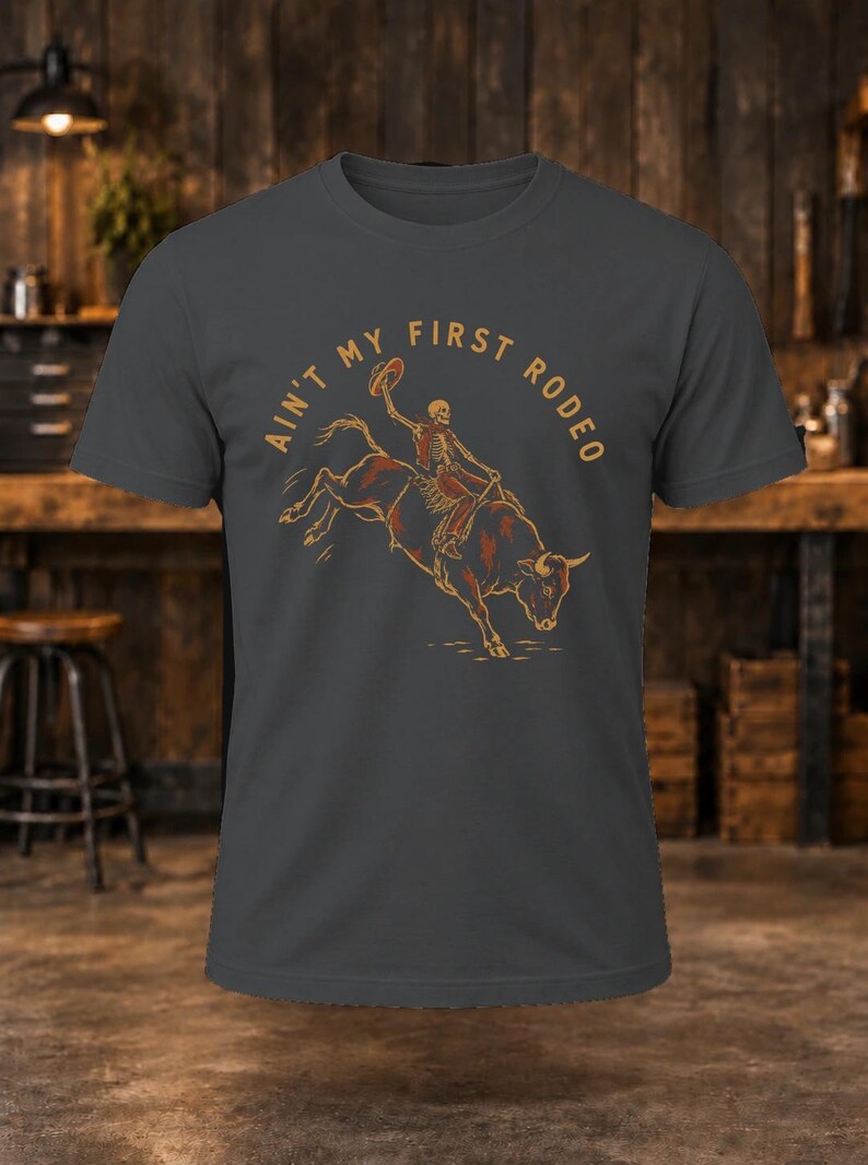 May include: A charcoal gray short-sleeve t-shirt featuring a graphic of a skeleton riding a bucking bull. The text "AIN'T MY FIRST RODEO" is printed in an arc above the skeleton in a tan color. The design is centered on the chest.