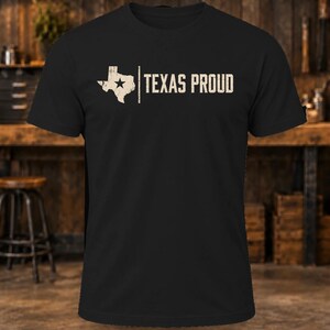 May include: A black short-sleeved t-shirt featuring a distressed white graphic of the state of Texas with a star inside, followed by a vertical line and the text "TEXAS PROUD" in a distressed white sans-serif font across the chest.