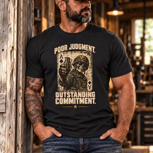 Puede incluir: Una camiseta negra de manga corta con un gr&aacute;fico de un soldado esqueleto con casco que sostiene una granada, enmarcado como una carta de as de picas. El texto dice POOR JUDGMENT y OUTSTANDING COMMITMENT con una peque&ntilde;a estrella.
