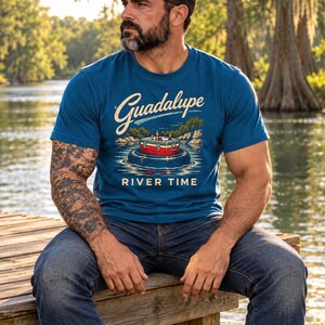 May include: A blue short-sleeve t-shirt featuring a graphic print of a red and white cooler floating in a black inner tube on water. The text Guadalupe is printed in a script font above the cooler, and the words RIVER TIME are printed below.