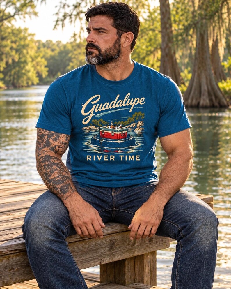 May include: A blue short-sleeve t-shirt featuring a graphic print of a red and white cooler floating in a black inner tube on water. The text Guadalupe is printed in a script font above the cooler, and the words RIVER TIME are printed below.