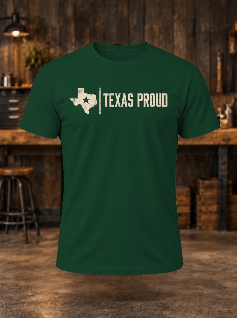 May include: A dark green short-sleeved t-shirt featuring a white graphic of the state of Texas with a star inside, followed by a vertical line and the text "TEXAS PROUD" in a distressed, block-style font across the chest.