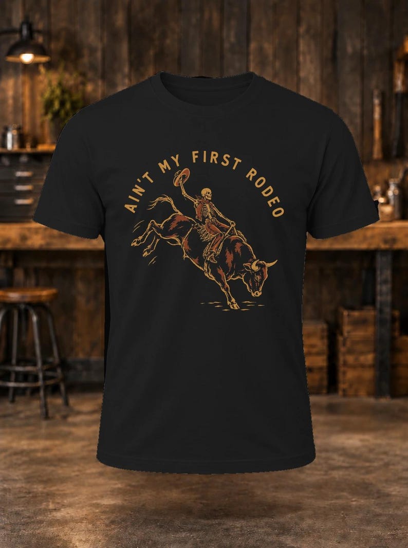 May include: A black short-sleeve t-shirt featuring a graphic of a skeleton riding a bucking bull. Above the illustration, the text reads AIN'T MY FIRST RODEO in a tan, arched font. The design is centered on the chest of the shirt.