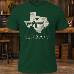 May include: A dark green short-sleeve t-shirt featuring a distressed white graphic of the state of Texas with a star and a rifle. Below the graphic, the text reads: TEXAS COME AND TAKE IT in a clean, white, sans-serif font.
