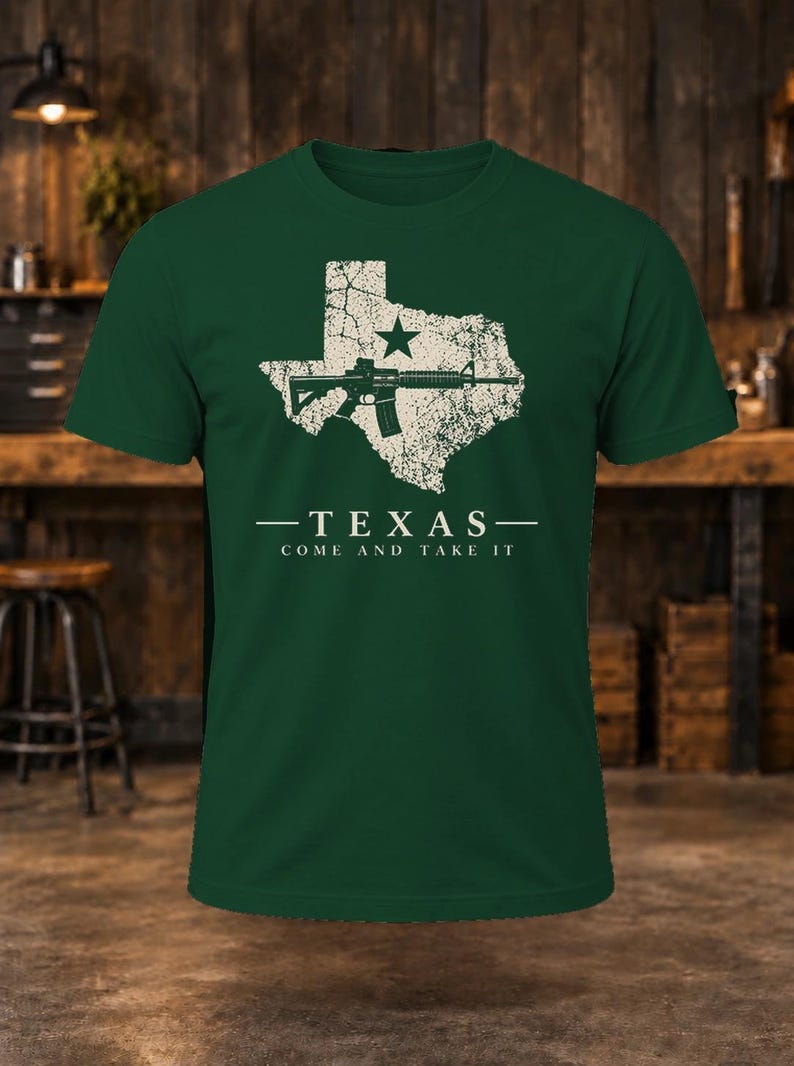 May include: A dark green short-sleeve t-shirt featuring a distressed white graphic of the state of Texas with a star and a rifle. Below the graphic, the text reads: TEXAS COME AND TAKE IT in a clean, white, sans-serif font.