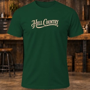 May include: A dark green short-sleeve t-shirt featuring the text "HILL COUNTRY" in a cream-colored, stylized font across the chest, with the word "TEXAS" printed in smaller letters underneath the main text on a curved line.