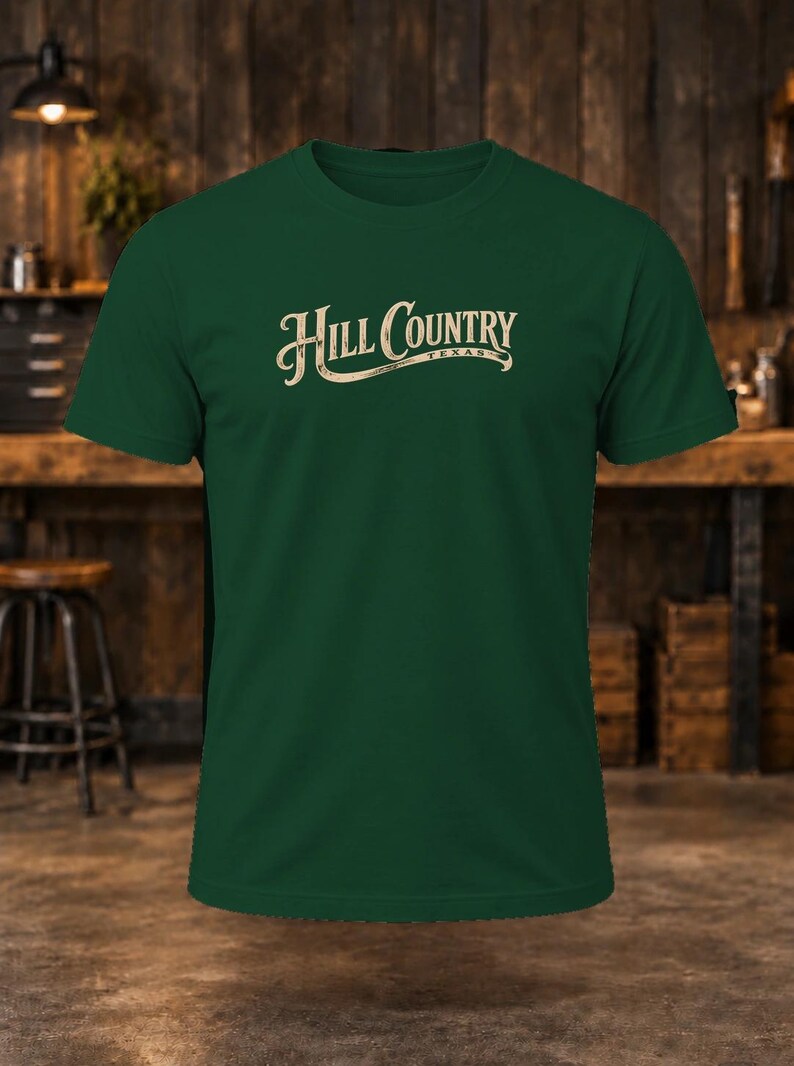 May include: A dark green short-sleeve t-shirt featuring the text "HILL COUNTRY" in a cream-colored, stylized font across the chest, with the word "TEXAS" printed in smaller letters underneath the main text on a curved line.