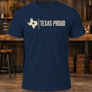 May include: A navy blue short-sleeve t-shirt featuring a distressed white graphic of the state of Texas with a star inside, followed by a vertical line and the text "TEXAS PROUD" in a bold, distressed white sans-serif font.