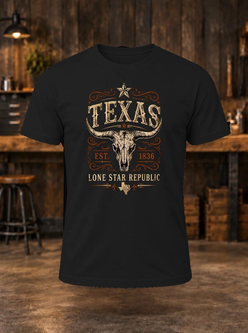 May include: A black short-sleeved t-shirt featuring a graphic print of a longhorn skull centred below the word TEXAS and a star. The design includes the text EST. 1836, LONE STAR REPUBLIC, and a small outline of the state of Texas.