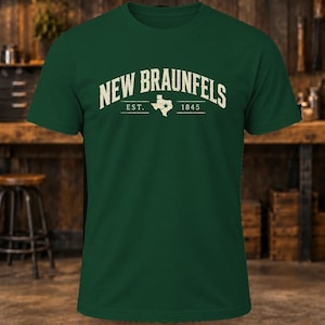 May include: A dark green short-sleeve t-shirt featuring the text "NEW BRAUNFELS" in a cream-colored arched font. Below the text is a graphic of the state of Texas with a star, flanked by the text "EST." and "1845" in a horizontal line.