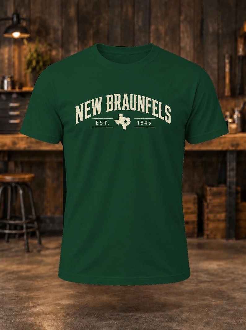 May include: A dark green short-sleeve t-shirt featuring the text "NEW BRAUNFELS" in a cream-colored arched font. Below the text is a graphic of the state of Texas with a star, flanked by the text "EST." and "1845" in a horizontal line.