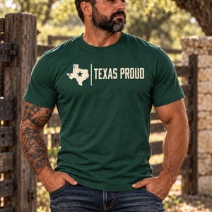 May include: A dark green short-sleeved crew neck t-shirt featuring a white distressed graphic of the state of Texas with a star inside, followed by the text "TEXAS PROUD" in a matching distressed white font across the chest.