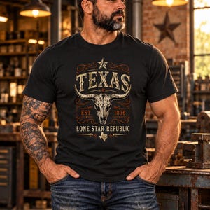 May include: A black short-sleeve t-shirt featuring a graphic print of a longhorn skull, a star, and the text "TEXAS EST. 1836 LONE STAR REPUBLIC" with a small map of Texas. The design is centred on the chest in shades of cream and orange.
