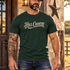 May include: A dark green short-sleeve crew neck t-shirt featuring the text Hill Country Texas printed in a light beige, stylized serif font across the chest. The shirt is paired with dark blue denim jeans and a tan baseball cap.