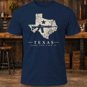 May include: A navy blue short-sleeve t-shirt featuring a distressed cream-colored graphic of the state of Texas with a star and a rifle. Below the graphic, the text reads: TEXAS - COME AND TAKE IT in a clean, sans-serif font.