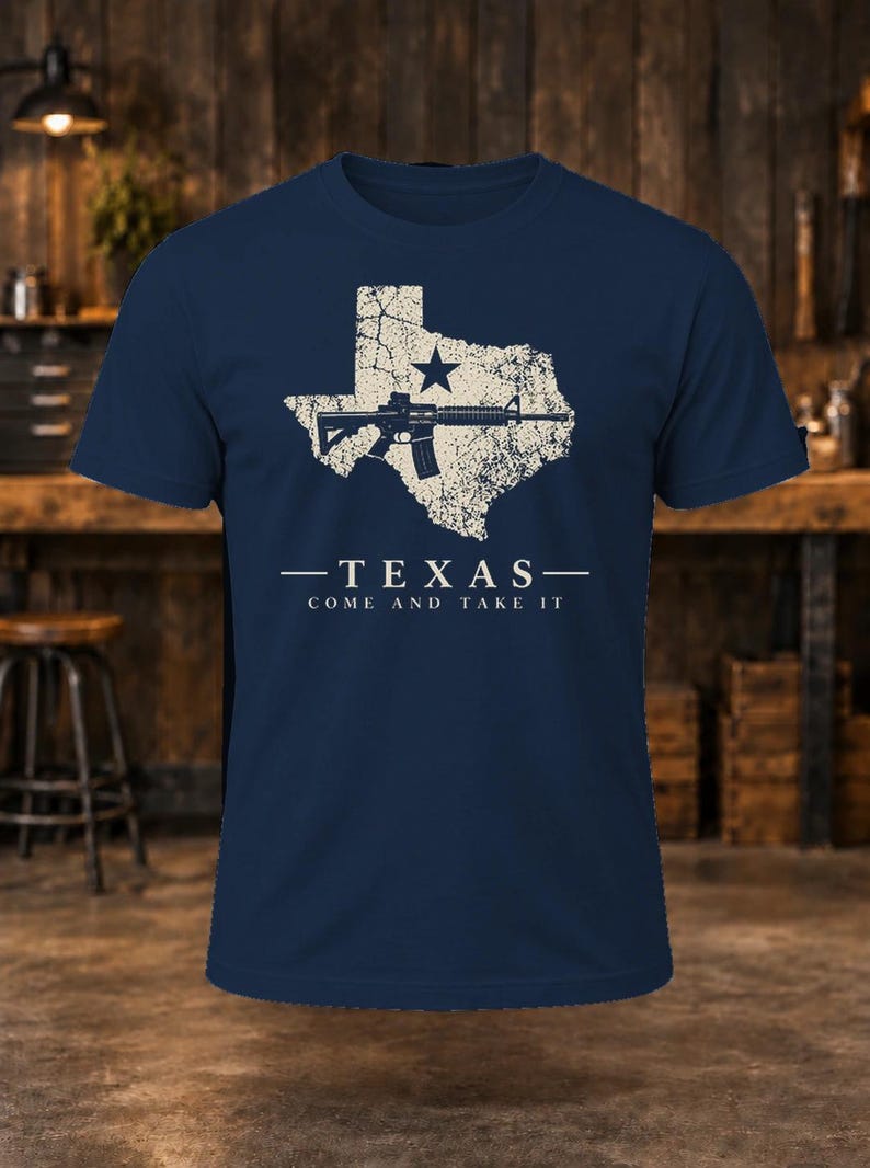 May include: A navy blue short-sleeve t-shirt featuring a distressed cream-colored graphic of the state of Texas with a star and a rifle. Below the graphic, the text reads: TEXAS - COME AND TAKE IT in a clean, sans-serif font.