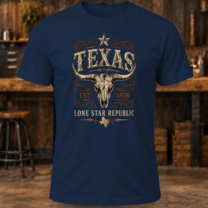 May include: A navy blue short-sleeve t-shirt featuring a graphic of a longhorn skull, a star, and the text "TEXAS EST. 1836 LONE STAR REPUBLIC" with a small map of Texas, all rendered in a distressed, weathered, off-white and orange colour palette.