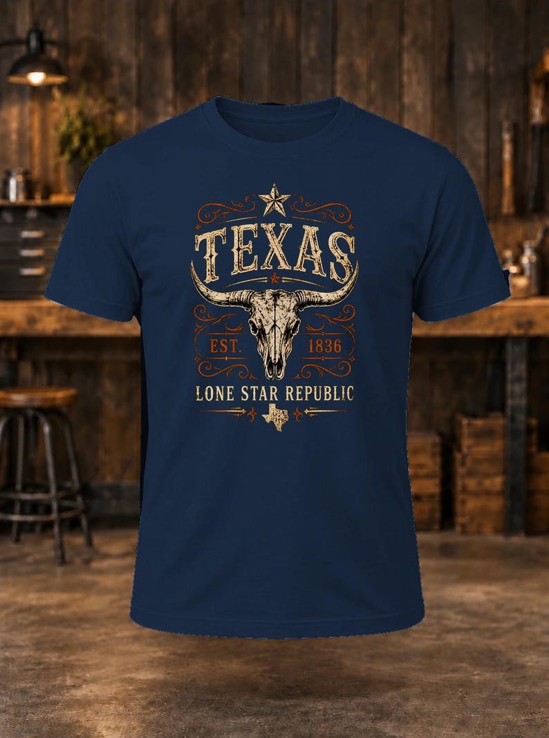 May include: A navy blue short-sleeve t-shirt featuring a graphic of a longhorn skull, a star, and the text "TEXAS EST. 1836 LONE STAR REPUBLIC" with a small map of Texas, all rendered in a distressed, weathered, off-white and orange colour palette.