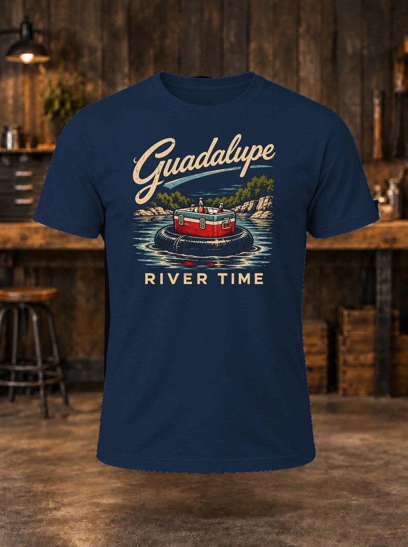 May include: A navy blue short-sleeve t-shirt featuring a graphic of a red and white cooler sitting on a black inner tube in a river. The text Guadalupe is printed in a cream script above, and RIVER TIME is printed in cream block letters below.