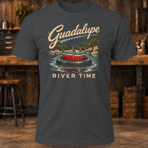 May include: A charcoal gray short-sleeve t-shirt featuring a graphic of a red and white cooler sitting on a black inner tube in a river. The text Guadalupe is printed in a script font above, and the words RIVER TIME are printed below.
