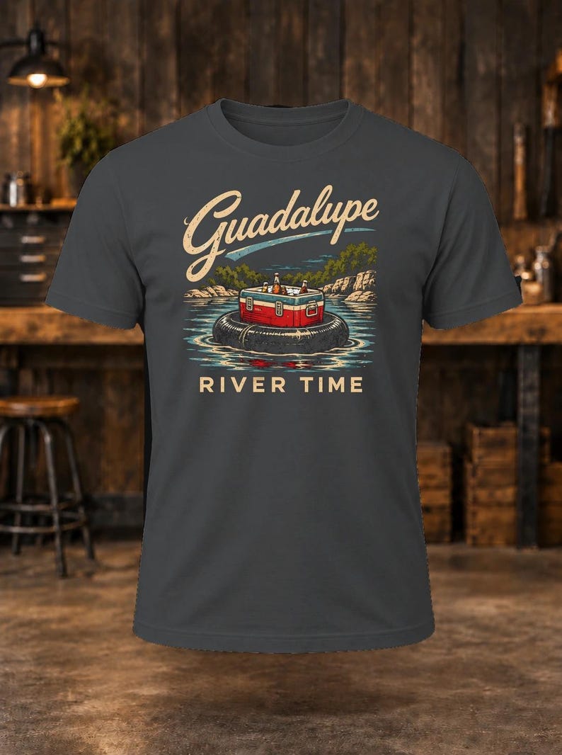 May include: A charcoal gray short-sleeve t-shirt featuring a graphic of a red and white cooler sitting on a black inner tube in a river. The text Guadalupe is printed in a script font above, and the words RIVER TIME are printed below.