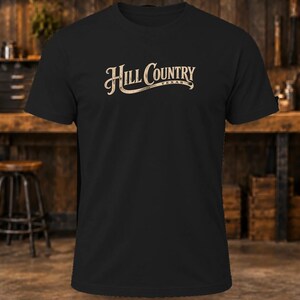 May include: A black short-sleeved t-shirt featuring the text Hill Country Texas printed in a light beige, stylized font across the chest. The design includes a decorative underline flourish beneath the word Country and the word Texas.