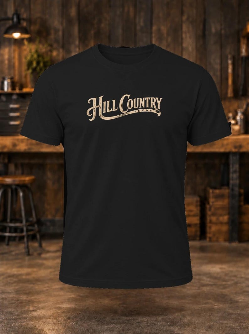 May include: A black short-sleeved t-shirt featuring the text Hill Country Texas printed in a light beige, stylized font across the chest. The design includes a decorative underline flourish beneath the word Country and the word Texas.