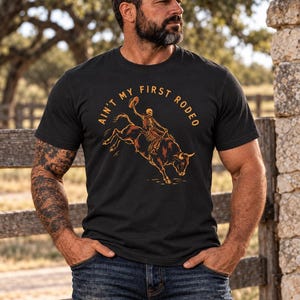 May include: A black short-sleeve t-shirt featuring a graphic print of a skeleton riding a bucking bull. The text "AIN'T MY FIRST RODEO" is printed in an arc above the illustration in a tan color. The design is centered on the chest.