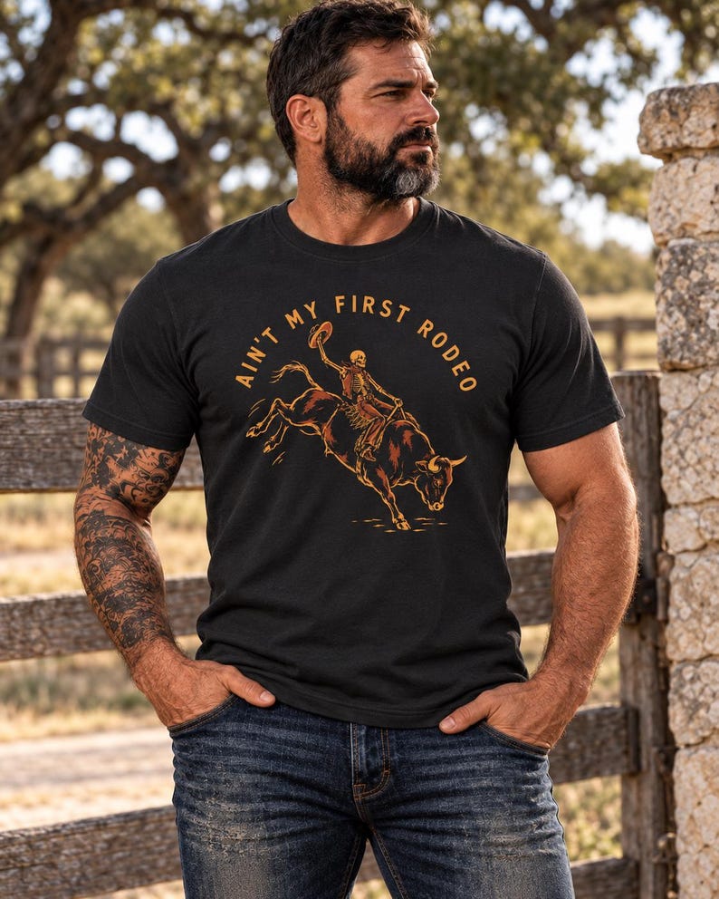 May include: A black short-sleeve t-shirt featuring a graphic print of a skeleton riding a bucking bull. The text "AIN'T MY FIRST RODEO" is printed in an arc above the illustration in a tan color. The design is centered on the chest.