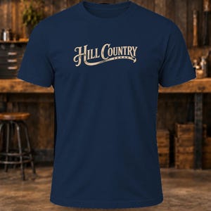 May include: A navy blue short-sleeve t-shirt featuring the text Hill Country Texas printed in a light beige, stylized script font across the center of the chest. The garment has a crew neckline and a standard fit.