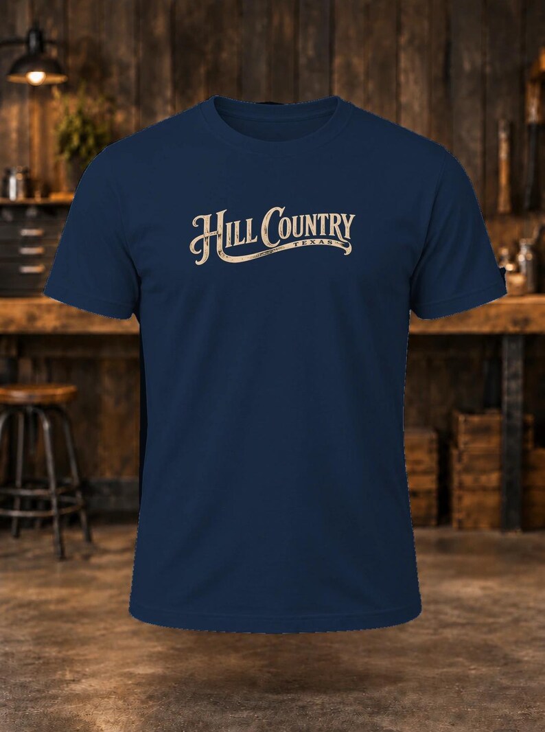 May include: A navy blue short-sleeve t-shirt featuring the text Hill Country Texas printed in a light beige, stylized script font across the center of the chest. The garment has a crew neckline and a standard fit.