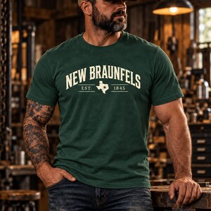 May include: A dark green short-sleeve t-shirt featuring the text NEW BRAUNFELS in a cream-colored arched font, with the abbreviation EST. 1845 and a graphic outline of the state of Texas with a star located in the center of the chest.
