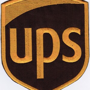 Puede incluir: Un parche bordado en forma de escudo con un fondo marrón oscuro, un borde amarillo dorado y una banda curva de color amarillo dorado en la parte superior. Las letras UPS están bordadas en una fuente amarilla dorada en el centro.