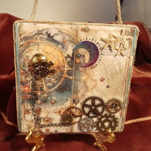 May include: A square wooden plaque with a vintage map design featuring a skull, gears, and a crescent moon. The plaque is decorated with gold and silver accents and has a distressed, aged look. The plaque is hanging from a twine rope and is displayed on a gold metal stand.