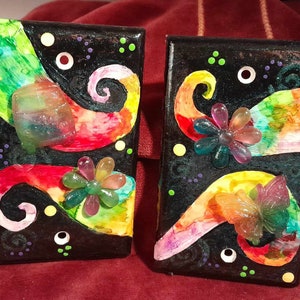 May include: Two small black square canvases with colorful abstract designs and 3D resin embellishments. The designs feature swirls, flowers, and butterflies in shades of pink, yellow, green, blue, and red.