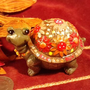 May include: A whimsical, hand-painted turtle figurine with a smiling face and a shell adorned with red, orange, and yellow floral designs. The turtle's body is a muted green, and it has small, detailed legs.