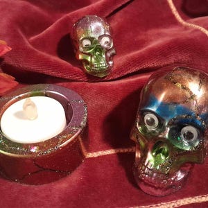 May include: Three iridescent skull candle holders with googly eyes. The candle holders are on a red velvet surface. One candle holder is holding a small white tea light.