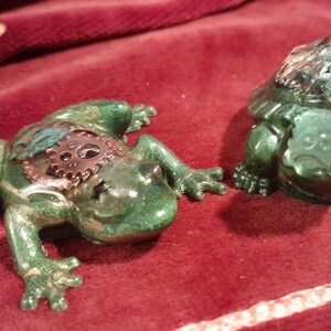May include: Two green decorative figurines: a frog with a gear-like design on its back and a turtle. The frog is lying down, and the turtle is upright. Both have a glittery finish and are set against a red background.