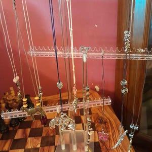 May include: A clear acrylic necklace display stand holding various necklaces with pendants. The necklaces feature different colored cords and chains, with pendants in silver, gold, and other colors. The stand is on a wooden surface.
