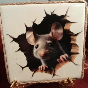 May include: Square ceramic tile featuring a realistic illustration of a mouse breaking through a wall. The mouse is gray and white with pink ears and nose. The tile is framed with a natural rope border and has gold-colored metal feet.