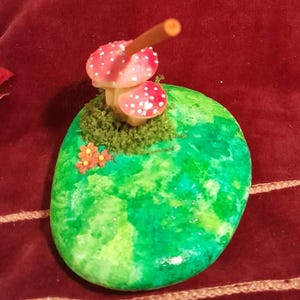May include: A painted stone with a green and yellow watercolor design. Two red and white spotted mushrooms are attached to the stone, along with a patch of green moss and small orange flowers. The stone is an oval shape.