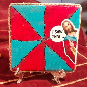 May include: A square, hand-painted artwork with a red and teal abstract design, framed with twine. A sticker of Jesus with the text "I SAW THAT..." is on the right side. The artwork is displayed on a decorative silver stand.