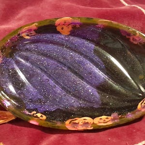 May include: A candy-shaped resin dish with a dark green and black exterior. The interior features a swirling pattern of purple glitter. Orange jack-o'-lanterns and purple accents decorate the edges, perfect for Halloween.