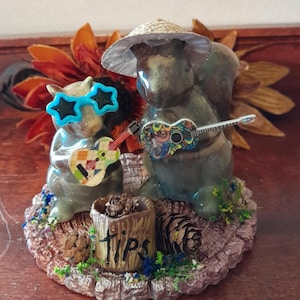 May include: A whimsical figurine featuring two squirrel musicians. One squirrel wears star-shaped sunglasses, the other a straw hat. Both squirrels hold colorful guitars. The base includes a "TIPS" sign and pine cones.