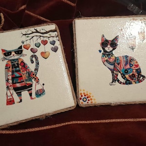 May include: Two square decorative tiles with cat illustrations. One cat wears sunglasses and a colorful outfit, holding bags. The other cat also wears sunglasses and a patterned outfit. Both tiles have rope edges and corner designs.
